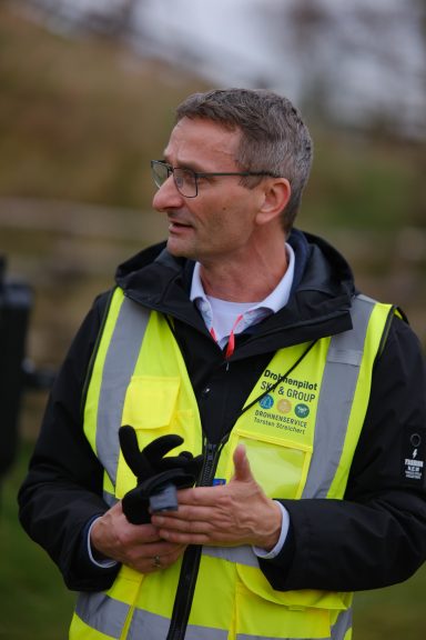 Mann in gelber Warnweste hält ein Mikrofon und spricht, im Freien.